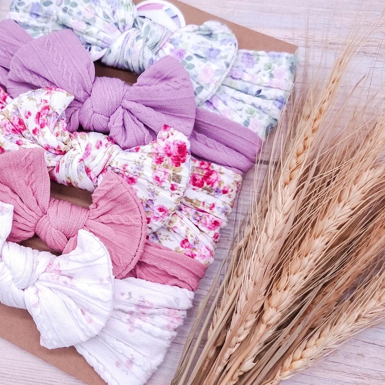 May include: A collection of five fabric headbands featuring knotted bows, arranged on a rectangular card. The headbands include solid mauve and pink cable-knit textures alongside white fabric with floral patterns in shades of pink, purple, and green.