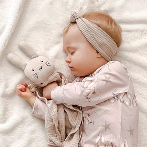 May include: A baby wearing a pink and white patterned onesie with a star design and a beige headband is sleeping while holding a beige bunny security blanket.