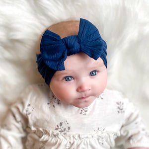 May include: A dark navy blue ribbed fabric headband featuring a large, centred bow detail. The headband is constructed from a soft, textured material that stretches to fit, showcasing a deep blue colour and a classic knotted bow design.