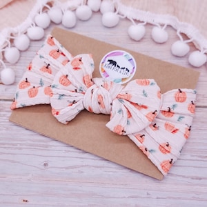 Könnte beinhalten: Ein weißes, geripptes Stoffstirnband mit orangefarbenem Kürbisdruck und einer mittigen Knoten-Schleife. Das Stirnband ist auf einer braunen Karte mit einem runden Aufkleber mit der Aufschrift Mama Bear & Cubs befestigt, umrandet von weißen Pompons.