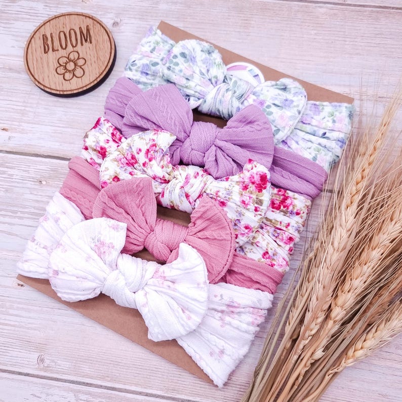 May include: A set of five fabric headbands with bows, featuring various floral patterns and solid colors including white, pink, and purple. The headbands are arranged on a rectangular card with a circular wooden tag labeled BLOOM.