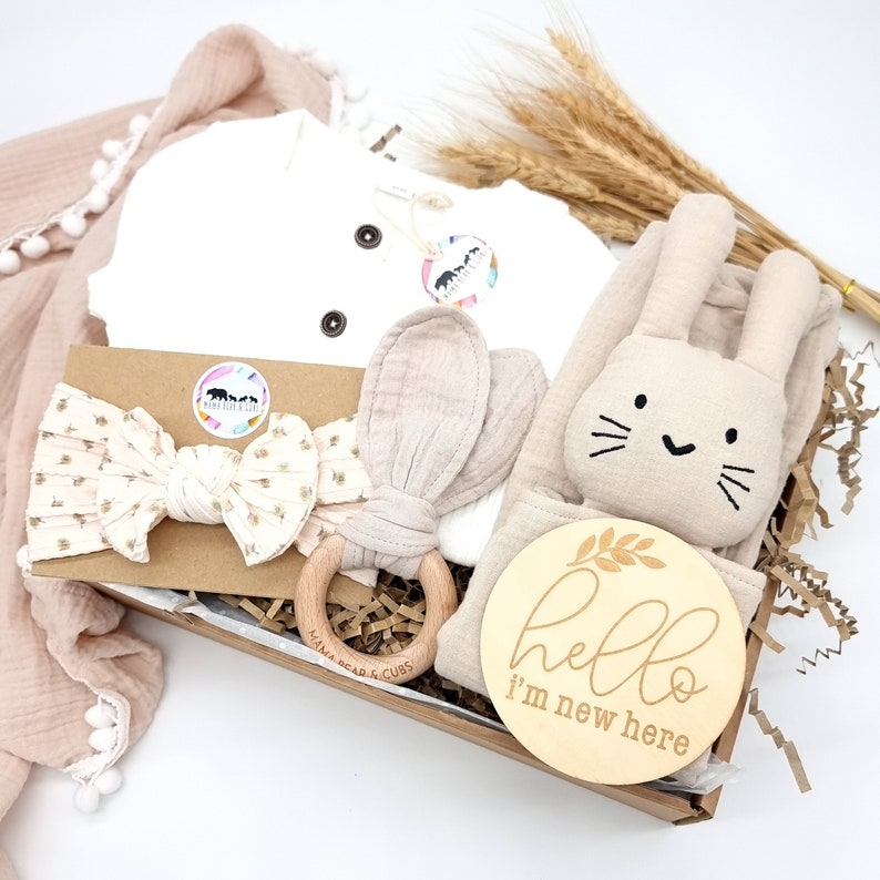 May include: A gift basket for a new baby with a white onesie, a wooden bunny rattle, a headband, a wooden ring, a beige muslin swaddle, and a wooden sign that says "hello i'm new here".