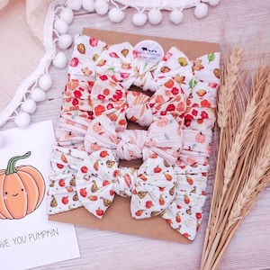 Könnte beinhalten: Vier weiße Stoffstirnbänder mit geknoteten Schleifen, jedes mit einem anderen herbstlichen Aufdruck, darunter bunte Herbstblätter, orangefarbene Kürbisse und kleine Zierkürbisse, präsentiert auf einer braunen Karte mit Kürbis-Illustration.