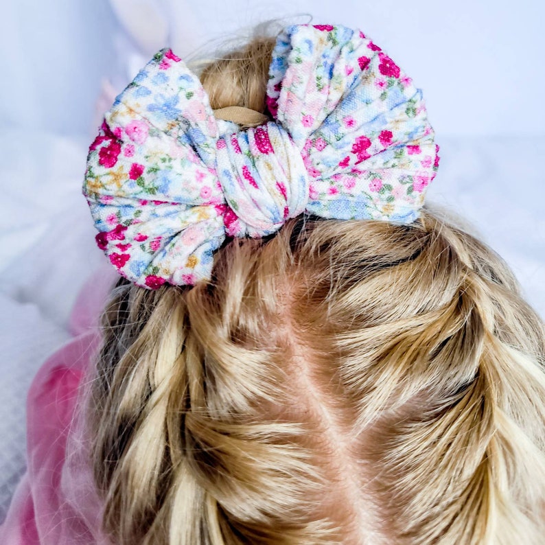 May include: A large hair bow featuring a white fabric base with a dense floral pattern in shades of bright pink, light blue, and green. The bow has a gathered, textured appearance and is secured to a tan elastic band, resting atop blonde hair.