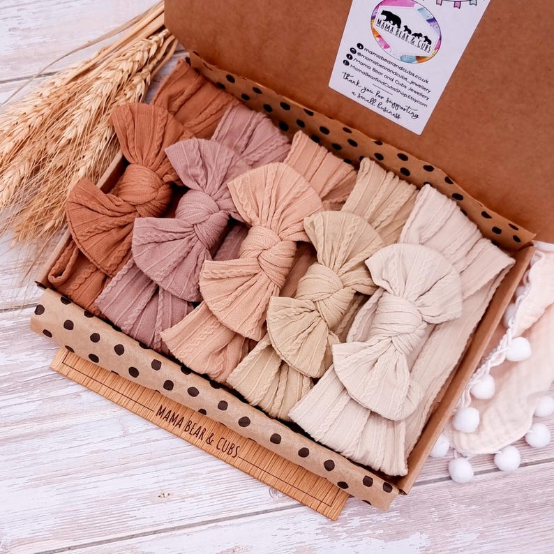 K&ouml;nnte beinhalten: Ein Set aus f&uuml;nf geknoten Haarb&auml;ndern in verschiedenen neutralen Farben, darunter Beige, Braun und Tan, sind in einer Schachtel mit schwarzen Punkten angeordnet. Die Schachtel ist mit braunem Papier ausgekleidet und hat den Text "Mama Bear & Cubs" auf der Unterseite.