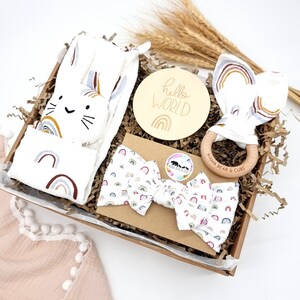 May include: A gift basket filled with baby items, including a white blanket with rainbow designs, a wooden teether with the text "Mama Bear & Cubs", a white headband with rainbow designs, a wooden sign with the text "hello world" and a rainbow design, and a white stuffed animal with a cat face and rainbow designs.