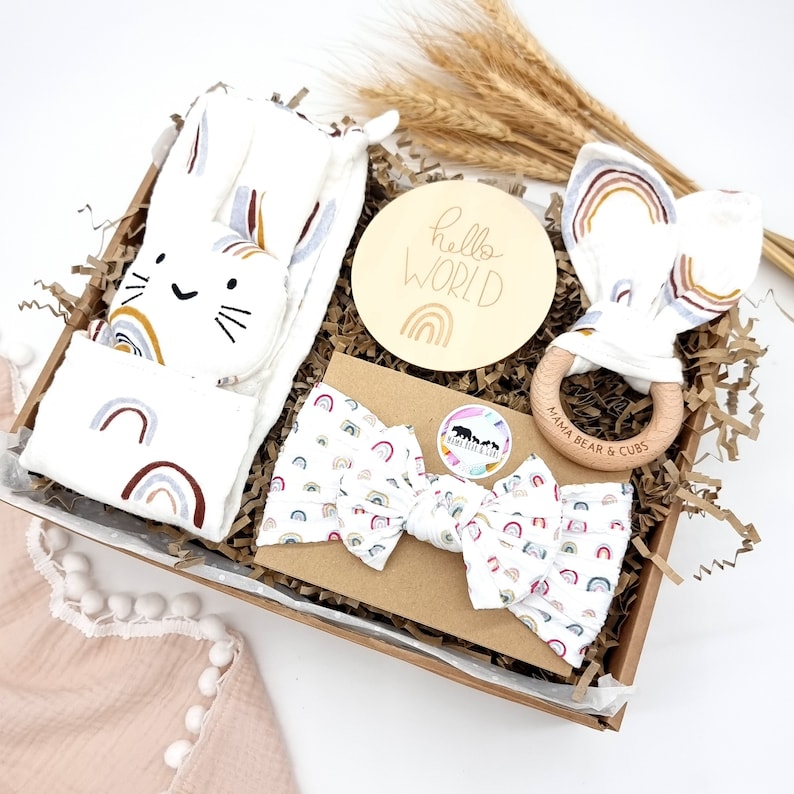 May include: A gift basket filled with baby items, including a white blanket with rainbow designs, a wooden teether with the text "Mama Bear & Cubs", a white headband with rainbow designs, a wooden sign with the text "hello world" and a rainbow design, and a white stuffed animal with a cat face and rainbow designs.