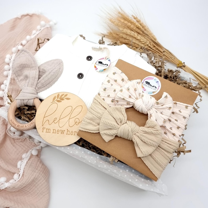 Puede incluir: Un juego de regalo para beb&eacute;s con un body blanco, diademas, un sonajero de madera y un letrero que dice "hello I'm new here". El juego incluye una manta suave de color rosa rubor con ribete de pomp&oacute;n y un sonajero de madera.