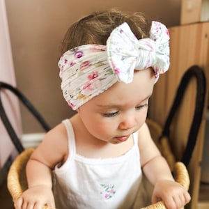 May include: A white fabric headband featuring a colorful floral pattern with pink, purple, and yellow flowers. The headband has a large, knotted bow detail at the center and a soft, ribbed texture. It is worn around the head of a young child.