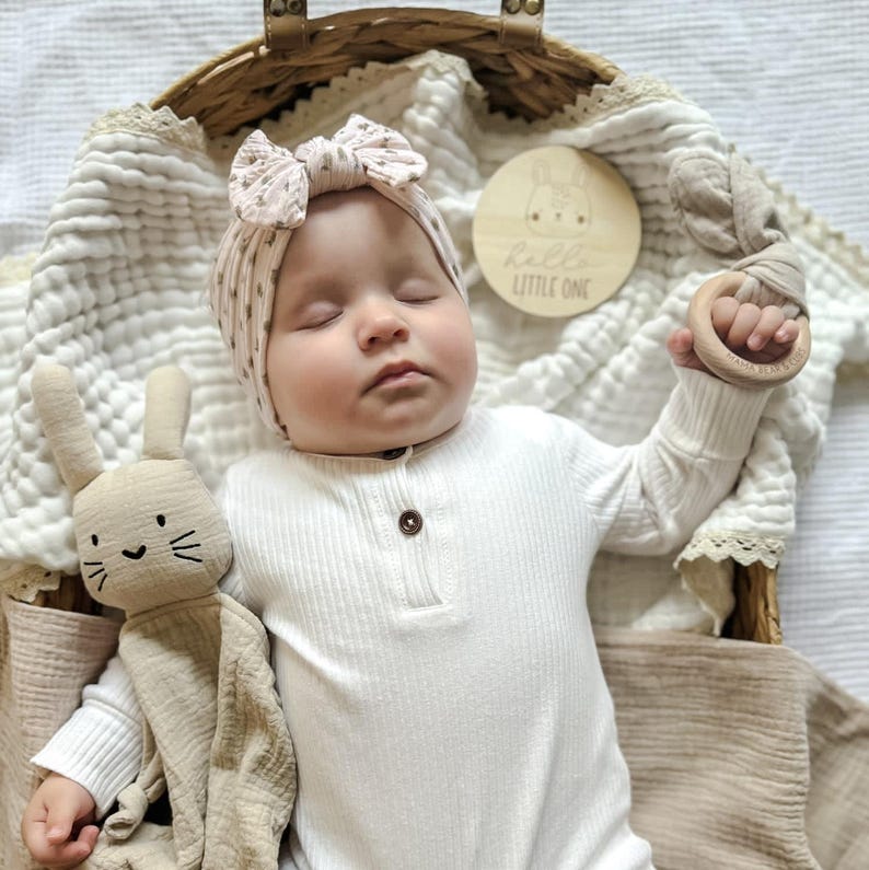 Puede incluir: Un beb&eacute; dormido con un body acanalado color crema y una diadema con lazo estampado. El beb&eacute; est&aacute; en una cesta tejida con un conejito de peluche y un mordedor de madera. Tambi&eacute;n hay un disco de madera con el texto "Hello Little One".