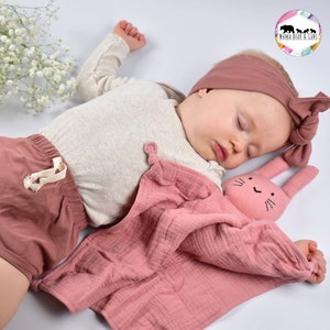 May include: A pink muslin baby blanket with a bunny attached. The blanket is soft and cuddly, perfect for a baby's comfort. The bunny has a simple black embroidered face.