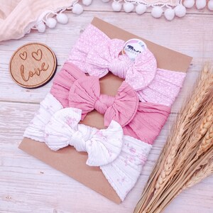 May include: Three pink and white patterned headbands with bows. The headbands are arranged on a brown background with a wooden sign that says "Love" in white letters.
