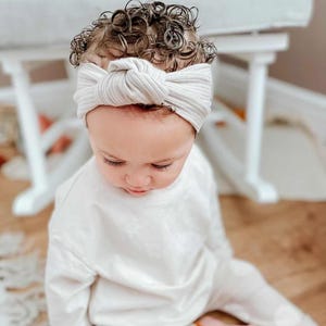 May include: A cream-coloured ribbed fabric headband featuring a central knot detail. The headband is worn across the forehead, contrasting with dark, curly hair. The material has a soft, textured appearance with visible vertical ribbing lines.
