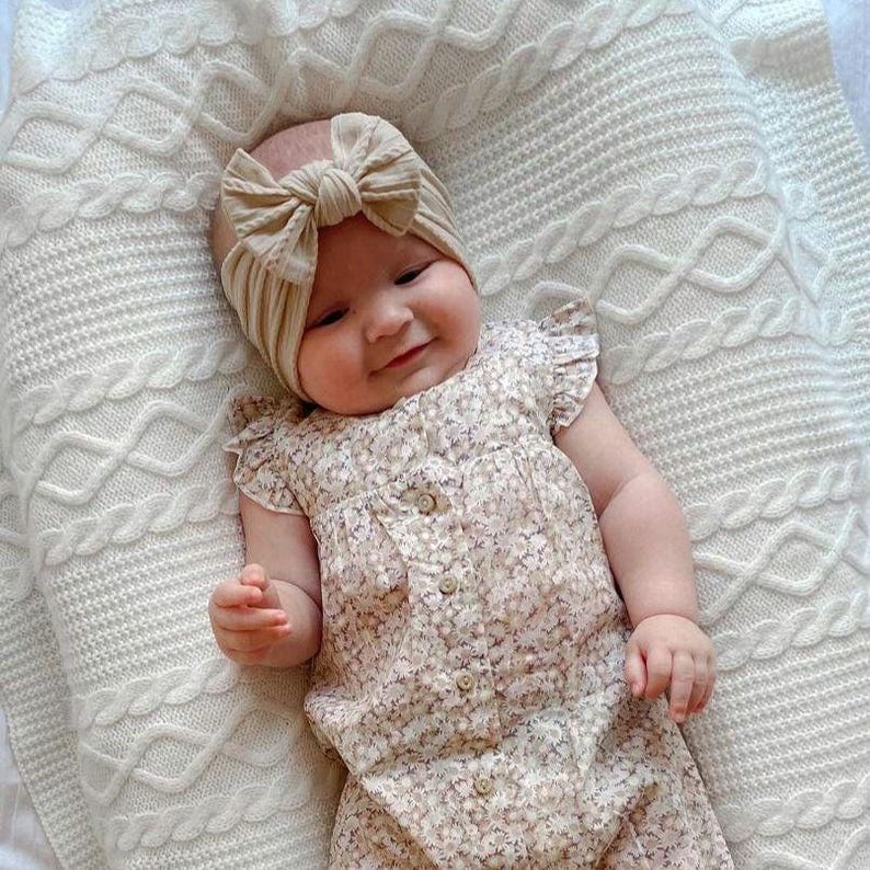 K&ouml;nnte beinhalten: Ein Baby tr&auml;gt einen floralen Strampler und ein beigefarbenes, gebundenes Stirnband. Das Baby liegt auf einer wei&szlig;en Strickdecke.