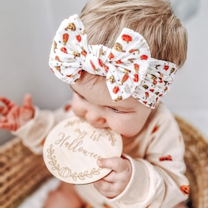 Peut inclure: Un bandeau en tissu blanc orné d'un motif répétitif de petites citrouilles et courges orange, porté avec un disque en bois rond gravé du texte "my 1st Halloween" entouré d'une bordure décorative de feuilles et de citrouilles.