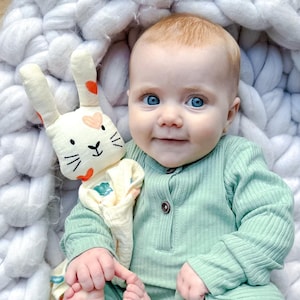May include: A baby in a light green long-sleeved onesie is holding a white and orange bunny lovey with a heart pattern. The baby is sitting in a white knitted basket.
