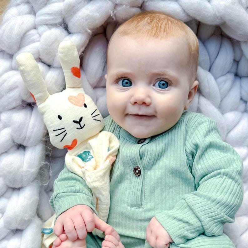 May include: A baby in a light green long-sleeved onesie is holding a white and orange bunny lovey with a heart pattern. The baby is sitting in a white knitted basket.