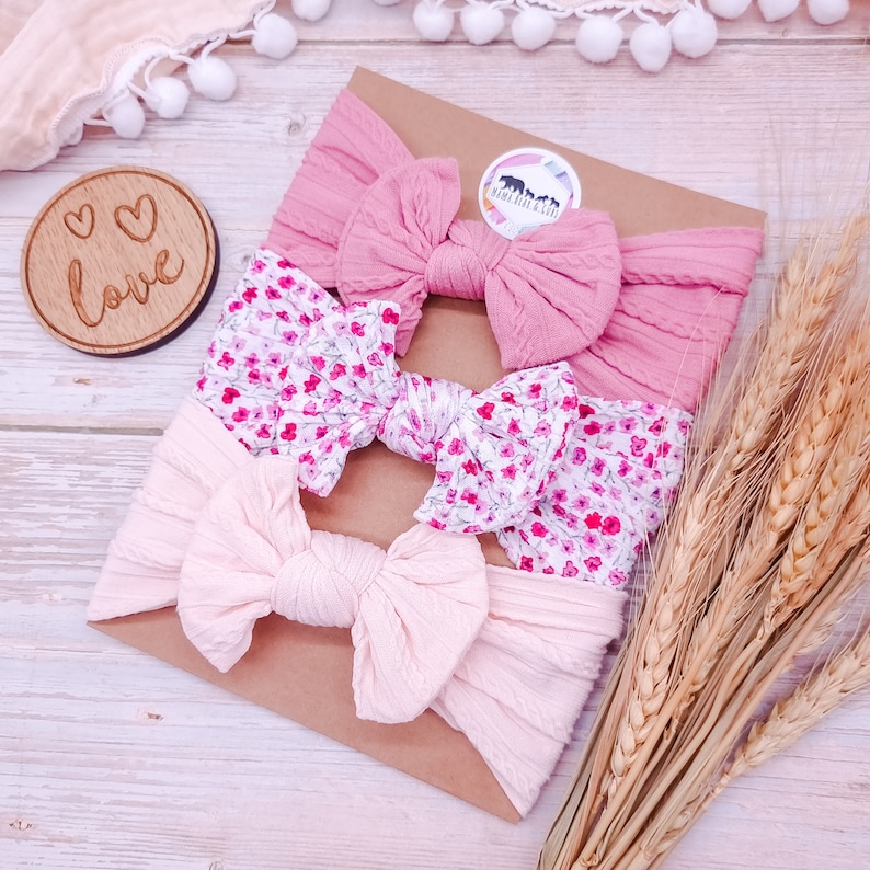 May include: Three pink and floral patterned headbands with bows. The headbands are arranged on a brown paper background with a small wooden sign that says "Love" in white lettering.