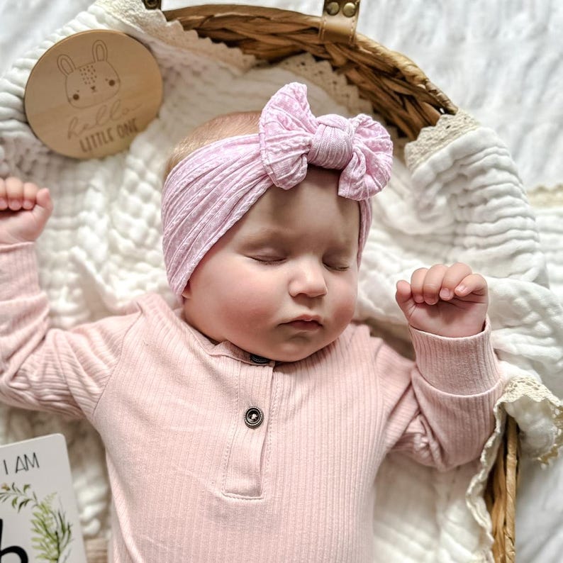 Puede incluir: Un beb&eacute; dormido con una camisa rosa de manga larga acanalada y una diadema con lazo rosa a juego. Un disco de madera con una ilustraci&oacute;n de conejito y el texto "hello LITTLE ONE" est&aacute; en el fondo. El beb&eacute; est&aacute; en una cesta tejida.