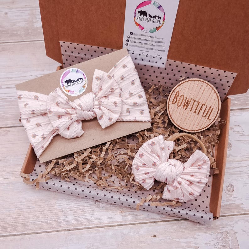 May include: A light pink ribbed fabric headband and a matching hair tie with a small floral pattern, presented in a cardboard gift box with brown paper filler. A round wooden disc with the text BOWTIFUL is included inside the box.