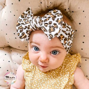 May include: A white fabric headband featuring a brown and black leopard print pattern with a large knotted bow detail at the center. The headband is worn on a head, paired with a mustard yellow sleeveless top with a small white floral print.