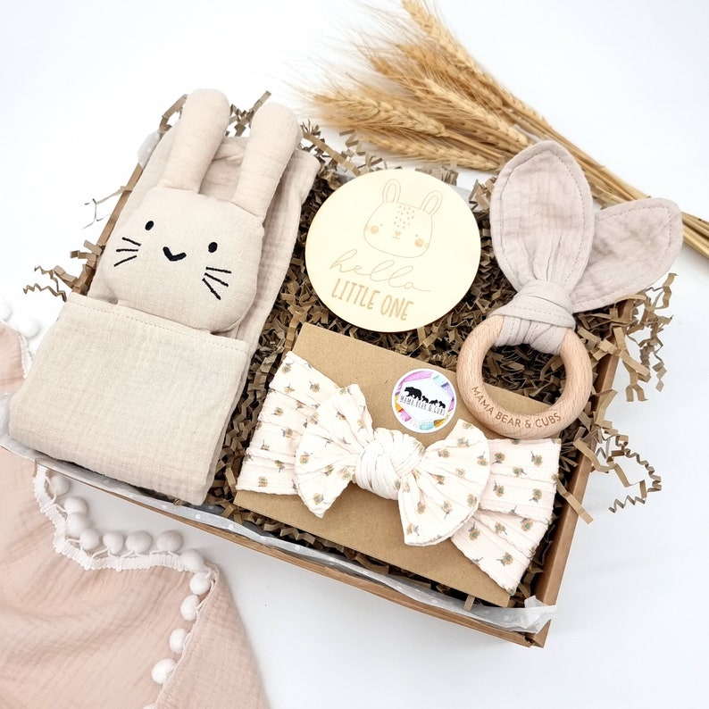 May include: A gift basket for a new baby with a bunny-shaped plush toy, a wooden ring with the text "Mama Bear & Cubs", a headband with a bow, and a wooden sign with the text "hello little one".