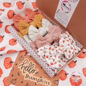 May include: A set of five fabric hair bows in a cardboard box, featuring solid colours in rust, mustard, cream, and dusty rose, plus one white bow with a pumpkin print. A card inside reads hello pumpkin, and a sticker shows Mama Bear & Cubs.