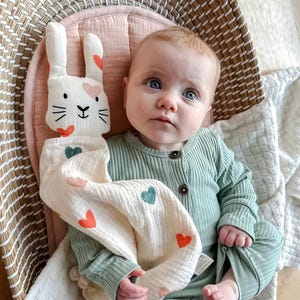May include: A baby in a green ribbed onesie holds a white muslin bunny lovey with pink and green hearts. The lovey has a white bunny face with black eyes and a black nose.