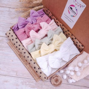 May include: A collection of seven ribbed fabric headbands with knotted bows in shades of purple, pink, sage green, yellow, and white, arranged in a brown cardboard gift box with polka dot trim and a label reading Mama Bear & Cubs.