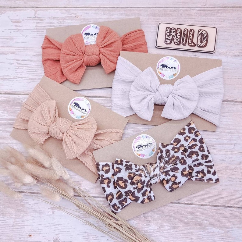 May include: Four headbands with large bows, each mounted on a cardboard display card. One is rust orange, one is cream, one is beige, and one features a brown and black leopard print. A wooden sign with the word WILD is included.