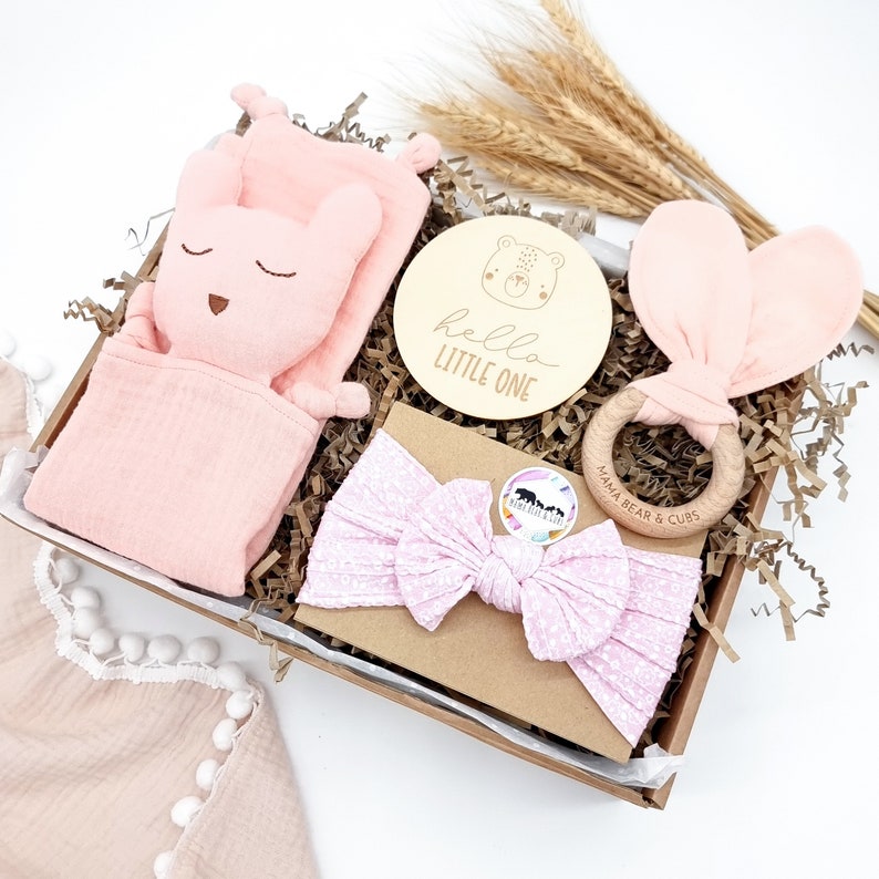May include: A pink and white baby gift box set with a plush bunny toy, a wooden ring, a headband, and a wooden sign that says "hello little one".