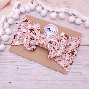 May include: A pink fabric headband featuring a repeating pattern of white ghosts, brown bats, and orange flowers. The headband has a knotted bow detail at the centre and is attached to a rectangular brown card with a circular logo reading Mama Bear & Cubs.