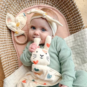 May include: A baby wearing a cream-colored knotted headband and a sage green ribbed long-sleeve shirt. The baby holds a cream-colored muslin security blanket with a rabbit head and heart patterns, alongside a wooden teething ring with bunny ears.