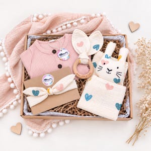 May include: A gift box containing a ribbed pink long-sleeve shirt with two buttons, a wooden teething ring with cream fabric bunny ears, a cream headband with heart prints, and a cream fabric square featuring a bunny face and heart patterns.