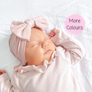 Könnte beinhalten: Ein hellrosa geripptes Langarmshirt mit Rüschenkragen und ein passendes geripptes Stirnband mit einer großen Schleife. Das Bild enthält eine kreisförmige rosa Grafik mit dem Text More Colours, die darauf hinweist, dass weitere Farben erhältlich sind.