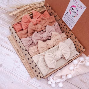 Könnte beinhalten: Ein Set aus fünf weichen, gerippten Stoffstirnbändern mit großen Schleifen, angeordnet in einem Farbverlauf von Terrakotta bis Creme. Die Stirnbänder sind in einem braunen Pappkarton mit schwarzen Punkten und einem Mama Bear & Cubs Aufkleber verpackt.