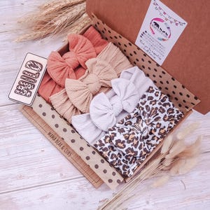 May include: A cardboard gift box containing four fabric headbands with bows. The headbands are in solid coral, beige, and white, plus one with a brown and black leopard print. A wooden tag reads WILD, and the box lid features a Mama Bear & Cubs logo.