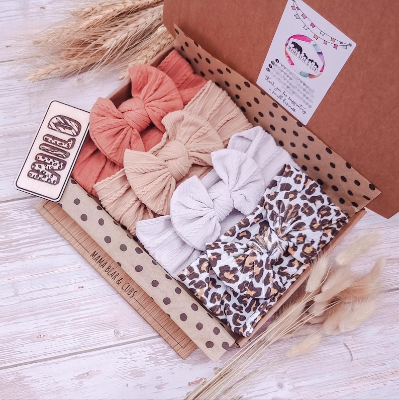 May include: A cardboard gift box containing four fabric headbands with bows. The headbands are in solid coral, beige, and white, plus one with a brown and black leopard print. A wooden tag reads WILD, and the box lid features a Mama Bear & Cubs logo.