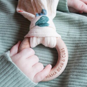 May include: A wooden teething ring with the engraved text MAMA BEAR & CUBS, featuring a soft, cream-colored fabric attachment decorated with blue and green leaf patterns, held against a sage green ribbed fabric background.