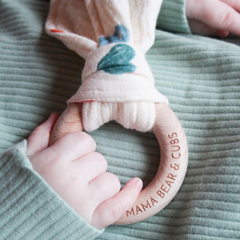 May include: A wooden teething ring with the engraved text MAMA BEAR & CUBS, featuring a soft, cream-colored fabric attachment decorated with blue and green leaf patterns, held against a sage green ribbed fabric background.