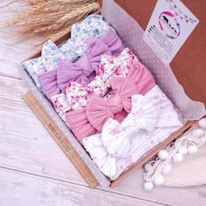 Könnte beinhalten: Eine Sammlung von fünf Stoffstirnbändern mit großen Schleifen, angeordnet in einer Pappschachtel. Die Stirnbänder zeigen verschiedene Blumenmuster und einfarbige Töne in Lavendel, Rosa und Weiß. Die Schachtel enthält den Text Mama Bear & Cubs.