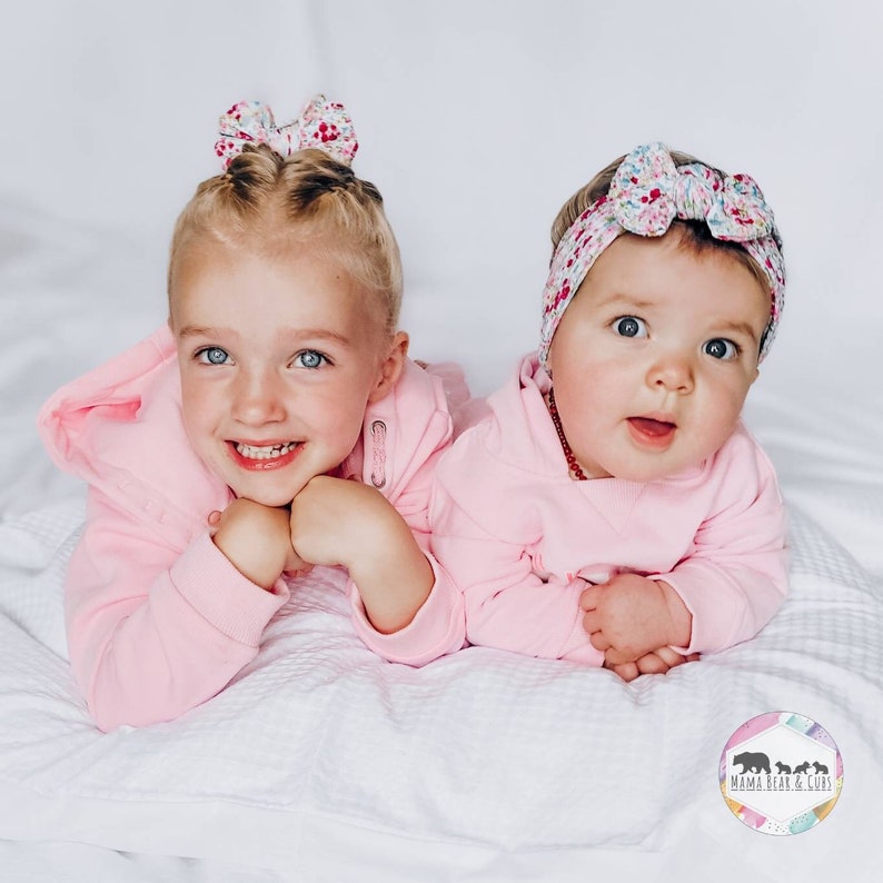 May include: Two light pink hooded sweatshirts paired with floral patterned hair accessories. One accessory is a small bow, and the other is a wide headband. A circular logo in the corner features the text Mama Bear & Curls with bear silhouettes.