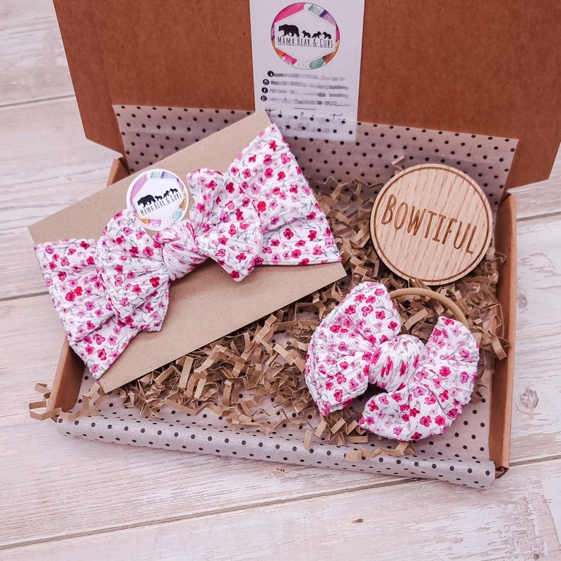 May include: A brown cardboard gift box containing a floral headband and a matching hair bow with a pink and white pattern. The box includes a circular wooden tag with the text BOWTIFUL and a branded card with the text MAMA BEAR & CUBS.