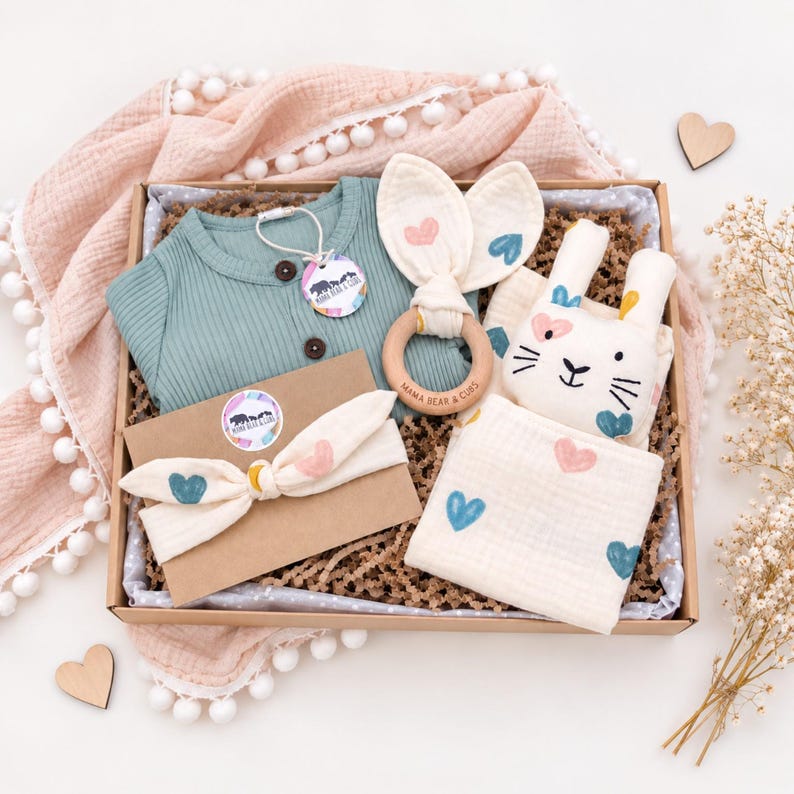 May include: A gift box containing a sage green ribbed long-sleeve shirt with a Mama Bear & Cubs tag, a wooden teething ring with cream fabric bunny ears, a cream headband with heart prints, and a cream muslin cloth with heart prints.