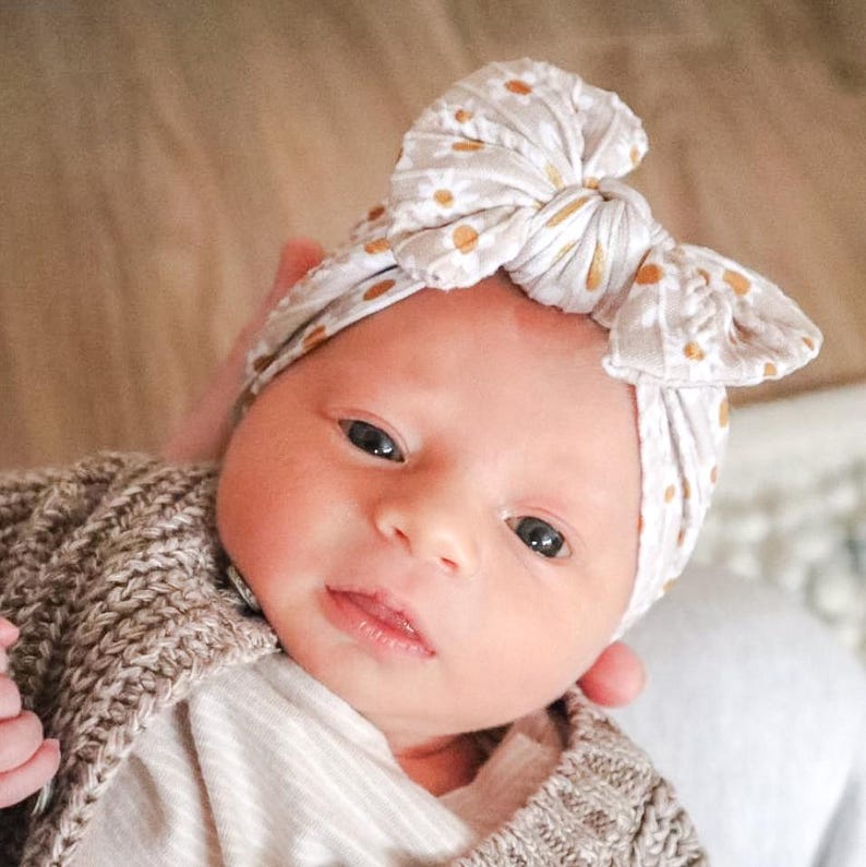 May include: A baby wearing a beige headband with a gold floral pattern and a beige knitted sweater. The headband is tied in a bow. The baby has dark eyes and is looking at the camera.