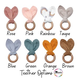 Puede incluir: Una colecci&oacute;n de mordedores para beb&eacute;s en varios colores, cada uno con un anillo de madera y orejas de conejo de tela. Los colores incluyen rosa, rosa, arco&iacute;ris, topo, azul, verde, naranja y marr&oacute;n. El texto "Teether Options" se muestra debajo.