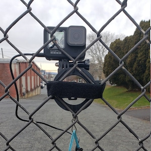 May include: A black GoPro camera mounted on a black metal bracket attached to a chain link fence. A black pouch with a blue drawstring is hanging from the bracket.