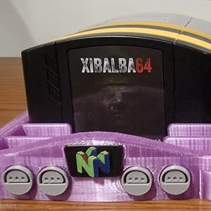 May include: A black and yellow video game cartridge labeled XIBALBA64, inserted into a purple 3D-printed plastic stand. The stand features a central N64-style logo emblem and four circular controller port cutouts along the front edge.