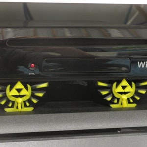 May include: A black Nintendo Wii U console with the text "Wii U" on the front. The console is sitting on two yellow 3D printed Triforce stands.