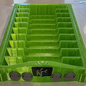May include: A lime green 3D-printed storage rack featuring ten vertical slots for game cartridges. The front panel displays a black rectangular emblem with the N64 logo and four circular gray controller ports integrated into the base structure.